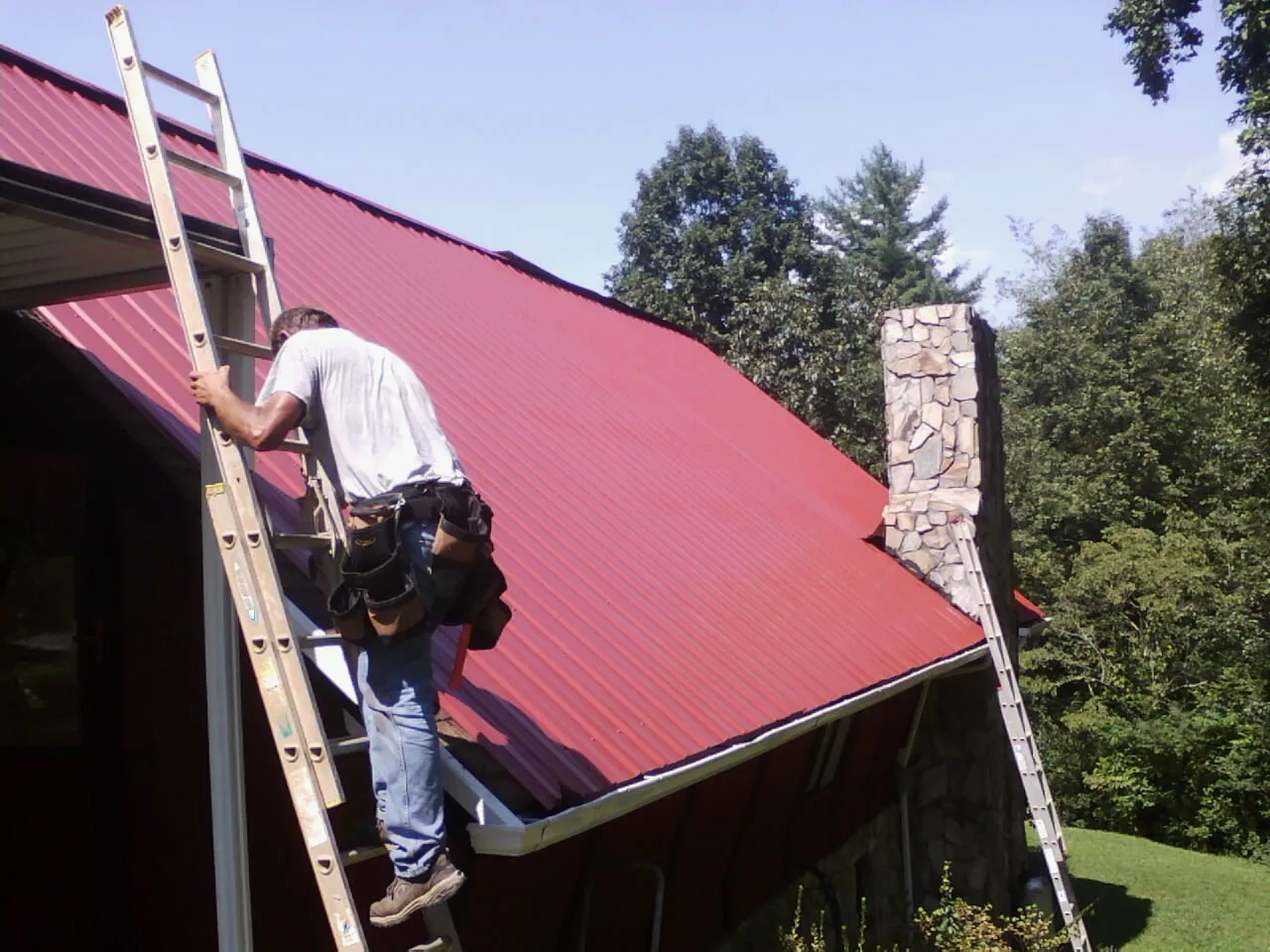 Roofer on a ladder beside a red metal roof and stone chimney — Soffit & Fascia Repair by Roofing Brothers in Tracy